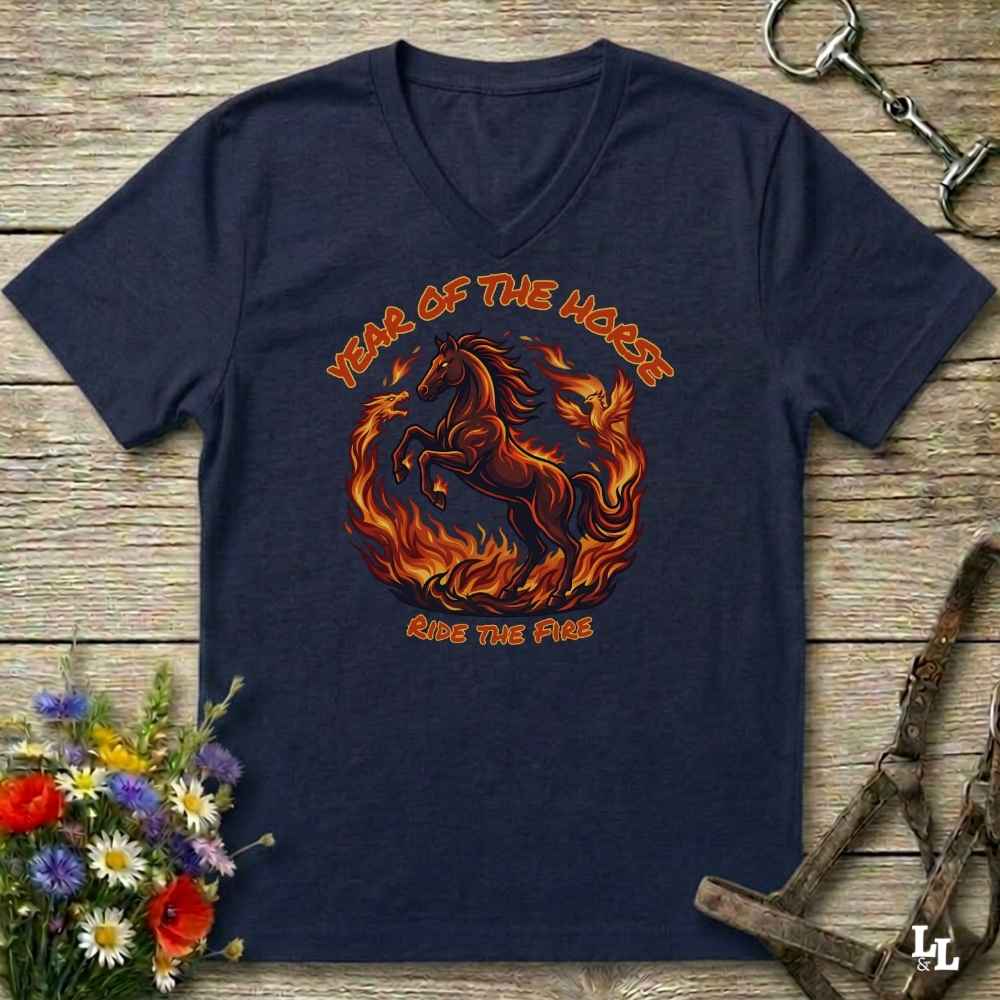 blue V-neck t-shirt laid flat on a rustic wooden surface, featuring a graphic of a rearing horse surrounded by flames with the text 'YEAR OF THE HORSE' and 'RIDE THE FIRE'. The shirt is styled with equestrian props including a leather bridle, horse bits, and a small bouquet of wildflowers.