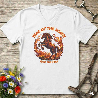 A white V-neck t-shirt laid flat on a rustic wooden surface, featuring a graphic of a rearing horse surrounded by flames with the text 'YEAR OF THE HORSE' and 'RIDE THE FIRE'. The shirt is styled with equestrian props including a leather bridle, horse bits, and a small bouquet of wildflowers.