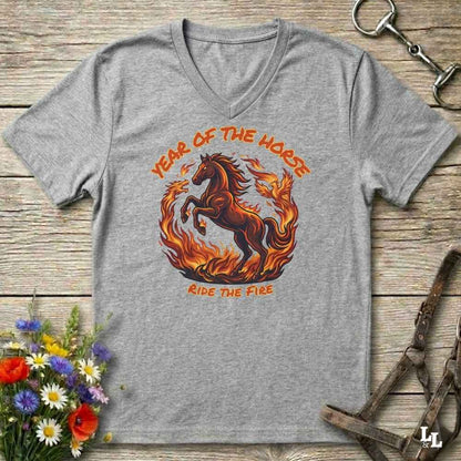 A heather gray V-neck t-shirt with a graphic of a rearing horse surrounded by flames, with the text 'YEAR OF THE HORSE' above and 'RIDE THE FIRE' below. The shirt is displayed flat on a rustic wooden background with decorative equestrian and floral props.