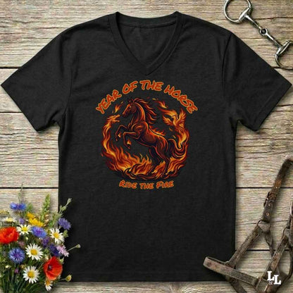 A black V-neck t-shirt with a graphic of a rearing horse engulfed in flames, with the text 'YEAR OF THE HORSE' above and 'RIDE THE FIRE' below, displayed on a rustic wooden background with horse tack and flowers.