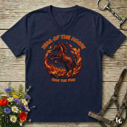 A navy blue V-neck t-shirt laid flat on a rustic wooden surface, featuring a graphic of a rearing horse surrounded by flames with the text 'YEAR OF THE HORSE' and 'RIDE THE FIRE'. The shirt is styled with equestrian accessories and flowers.