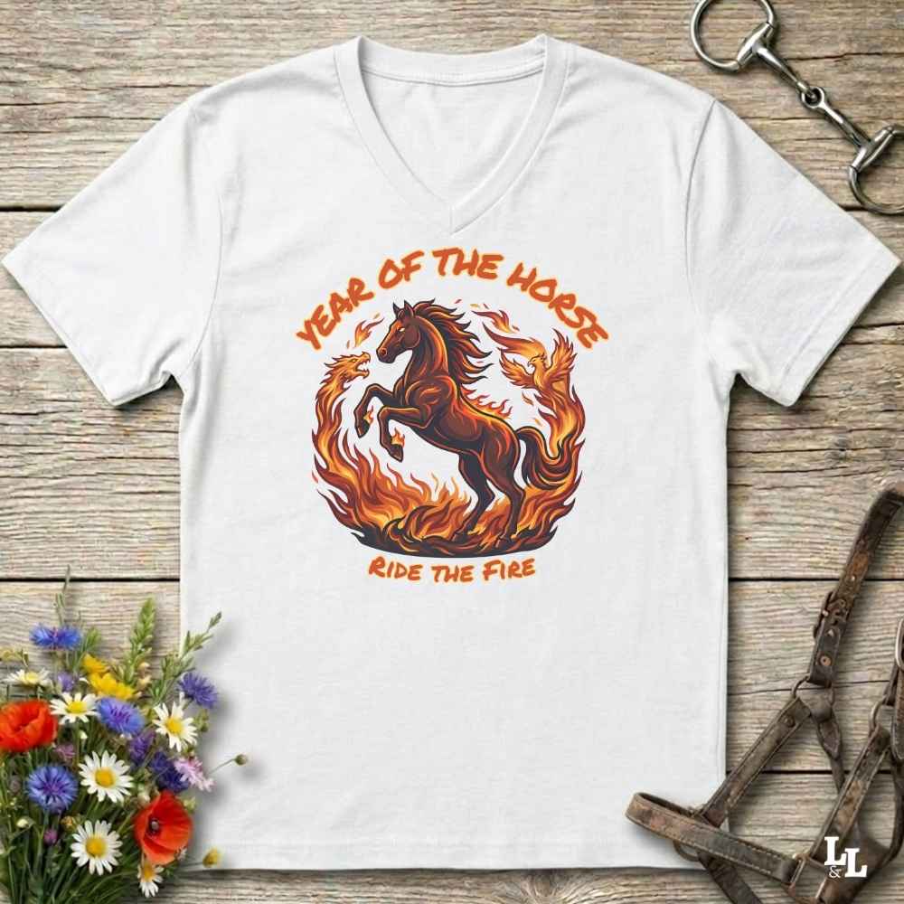A white V-neck t-shirt laid flat on a rustic wooden surface, featuring a graphic of a rearing horse surrounded by flames with the text 'YEAR OF THE HORSE' and 'RIDE THE FIRE'. The shirt is styled with equestrian props including a leather bridle, horse bits, and a small bouquet of wildflowers.