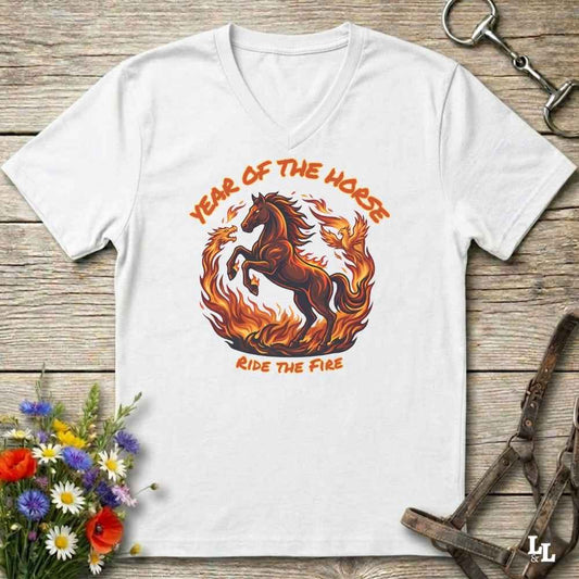 A white V-neck t-shirt laid flat on a rustic wooden surface, featuring a graphic of a rearing horse surrounded by flames with the text 'YEAR OF THE HORSE' and 'RIDE THE FIRE'. The shirt is styled with equestrian props including a leather bridle, horse bits, and a small bouquet of wildflowers.