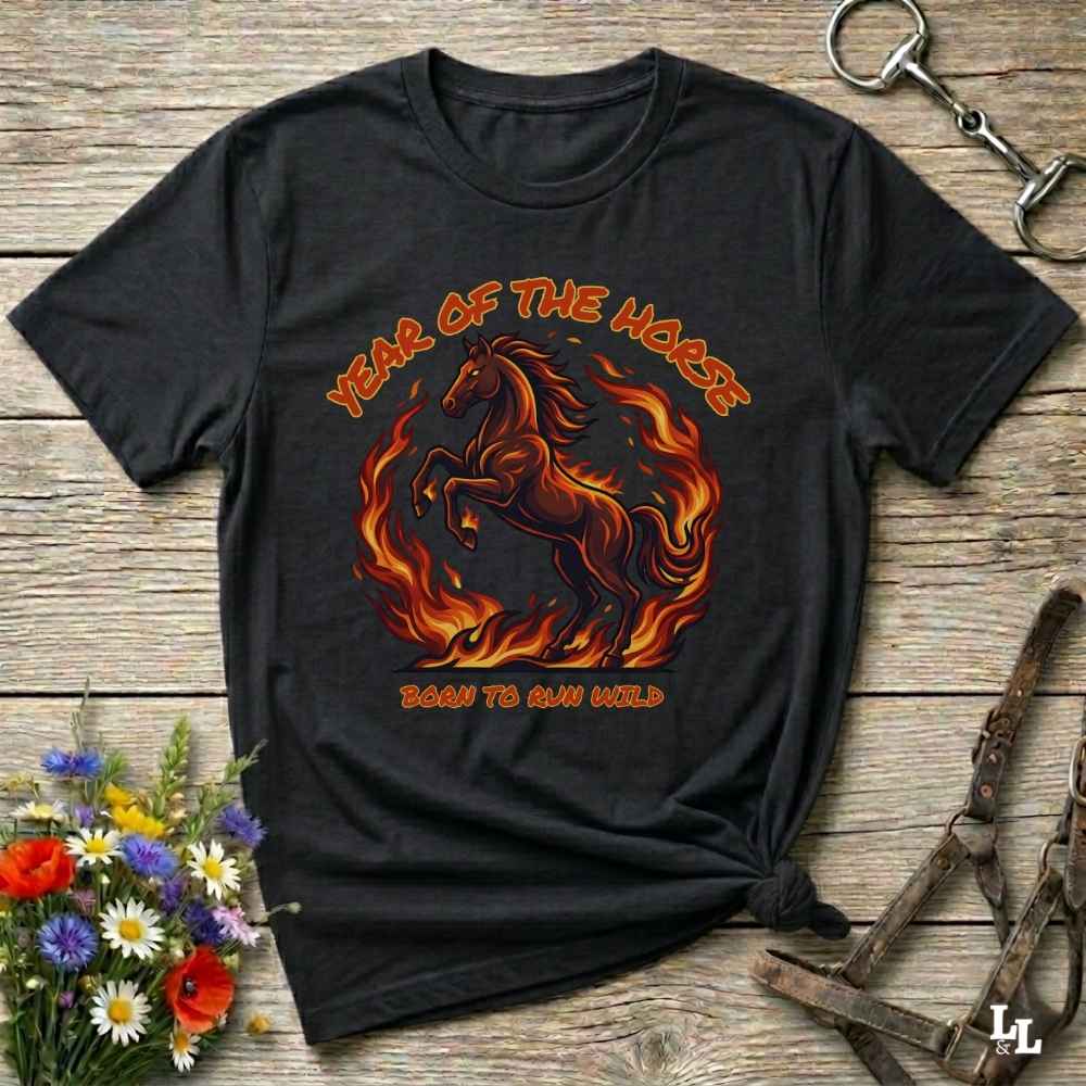 A black graphic t-shirt featuring a fiery red and orange horse design with the text 'YEAR OF THE HORSE' and 'BORN TO RUN FREE', laid flat on a rustic wooden background with decorative flowers and tack.