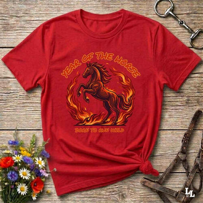 A red short-sleeve graphic T-shirt laid flat on a rustic wood surface, with a horse and flames printed on the front and the words 'YEAR OF THE HARE' and 'BORN TO RUN WILD.' Decorative flowers and horse tack are placed around it as styling props.
