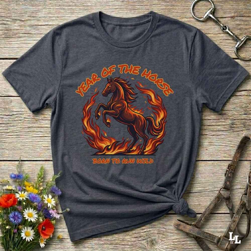 A dark heather gray graphic t-shirt laid flat on a rustic wooden surface. The shirt features a graphic of a rearing horse surrounded by flames with the text 'YEAR OF THE HORSE' and 'BORN TO RUN WILD'.