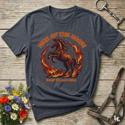 A dark heather gray graphic t-shirt laid flat on a rustic wooden surface. The shirt features a graphic of a rearing horse surrounded by flames with the text 'YEAR OF THE HORSE' and 'BORN TO RUN WILD'.