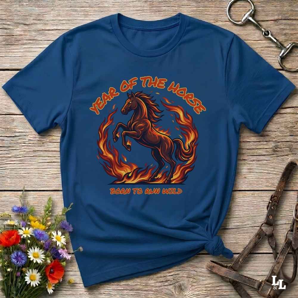 A navy blue graphic t-shirt laid flat on a rustic wooden surface, styled with a horse halter, flowers, and a horse tack. The shirt features a graphic of a rearing horse surrounded by flames with the text 'YEAR OF THE HORSE' and 'BORN TO RUN WILD'.