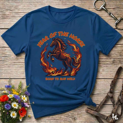 A navy blue graphic t-shirt laid flat on a rustic wooden surface, styled with a horse halter, flowers, and a horse tack. The shirt features a graphic of a rearing horse surrounded by flames with the text 'YEAR OF THE HORSE' and 'BORN TO RUN WILD'.