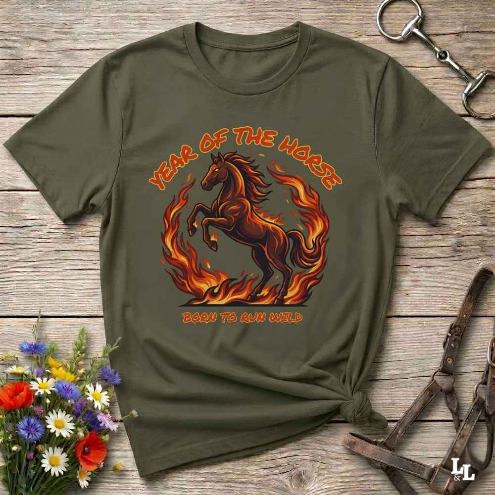 An olive green graphic t-shirt laid flat on a rustic wooden surface, styled with a horse halter, flowers, and a small knot at the bottom right. The t-shirt features a graphic of a fiery brown horse with the text 'YEAR OF THE HORSE' and 'BORN TO RUN FREE'.