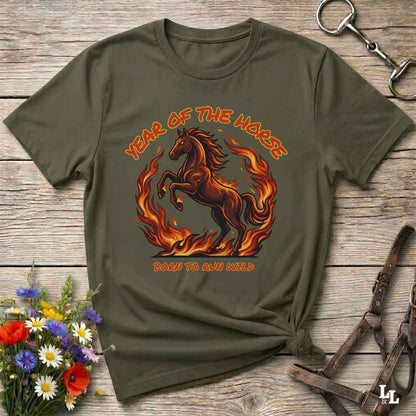 An olive green graphic t-shirt laid flat on a rustic wooden surface, styled with a horse halter, flowers, and a small knot at the bottom right. The t-shirt features a graphic of a fiery brown horse with the text 'YEAR OF THE HORSE' and 'BORN TO RUN FREE'.