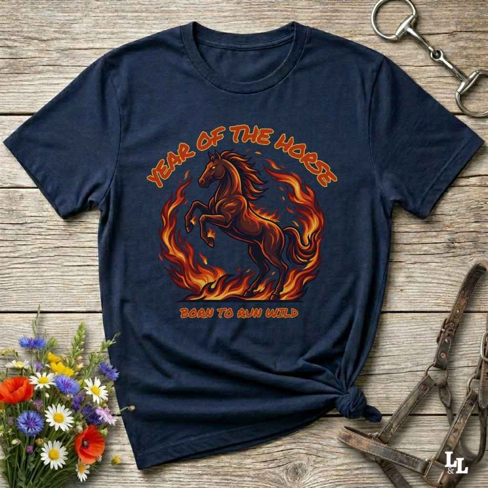 A navy blue graphic t-shirt featuring a fiery red and orange horse design with the text 'YEAR OF THE HORSE' and 'BORN TO RUN WILD', styled flat on a rustic wooden background with decorative flowers and horse tack.