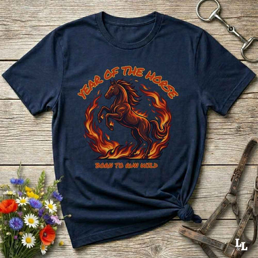 A navy blue graphic t-shirt featuring a fiery red and orange horse design with the text 'YEAR OF THE HORSE' and 'BORN TO RUN WILD', styled flat on a rustic wooden background with decorative flowers and horse tack.