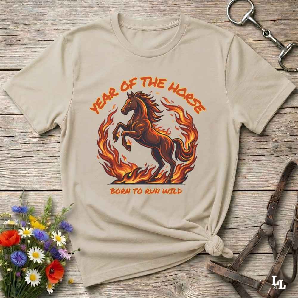 A beige graphic t-shirt featuring a brown horse running through orange flames with the text 'YEAR OF THE HORSE' and 'BORN TO RUN WILD'. The shirt is styled in a flat-lay on a rustic wooden background with equestrian accessories and flowers.