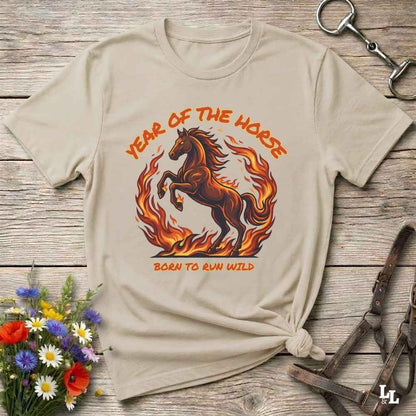 A beige graphic t-shirt featuring a brown horse running through orange flames with the text 'YEAR OF THE HORSE' and 'BORN TO RUN WILD'. The shirt is styled in a flat-lay on a rustic wooden background with equestrian accessories and flowers.