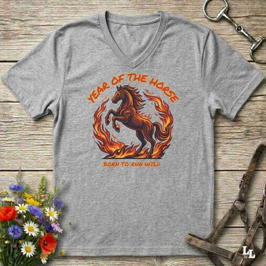 A heather gray V-neck t-shirt featuring a graphic of a brown horse running through orange flames, with the text 'YEAR OF THE HORSE' above and 'BORN TO RUN WILD' below. The shirt is displayed flat on a rustic wooden background with equestrian accessories and flowers.