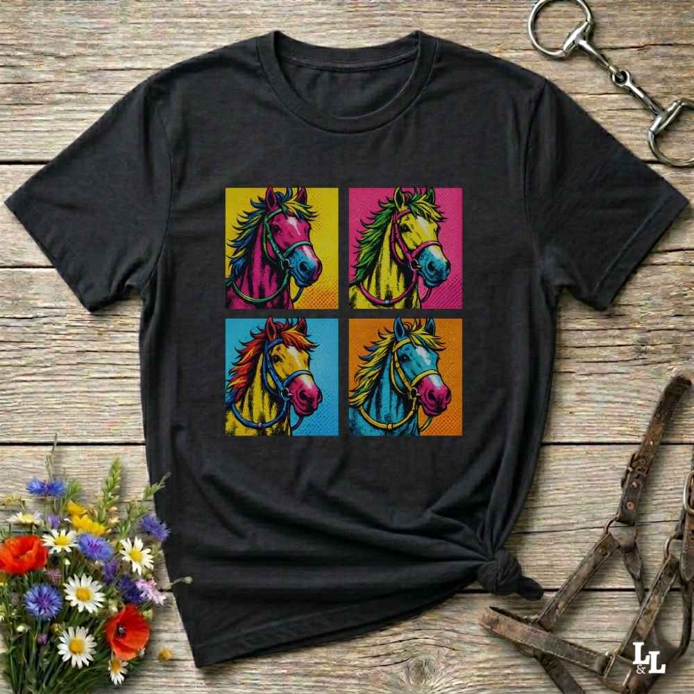 A black short-sleeve t-shirt with a colorful graphic print of four pop-art horses in a 2x2 grid, styled on a rustic wooden background with equestrian accessories and flowers.