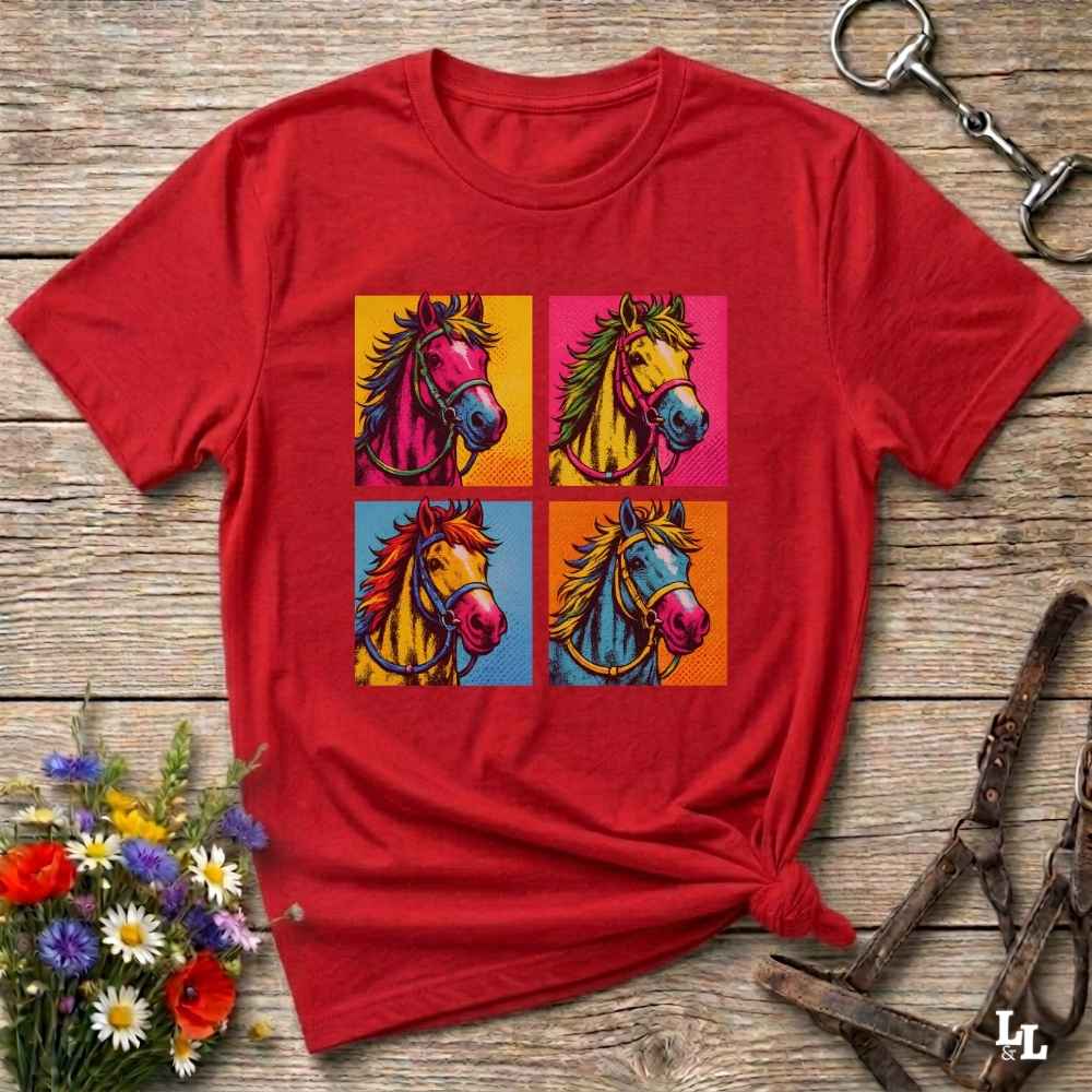 A red short-sleeve t-shirt with a colorful pop-art graphic of four horse heads in a 2x2 grid, styled on a rustic wooden background with equestrian accessories and flowers.