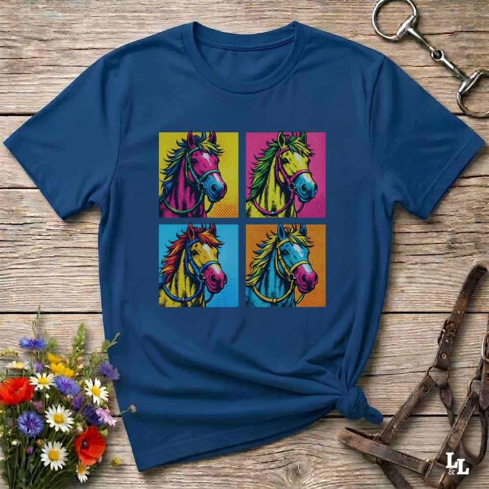 A navy blue short-sleeve t-shirt with a colorful pop-art graphic of four horse heads in a 2x2 grid, styled on a rustic wooden background with equestrian accessories and flowers.