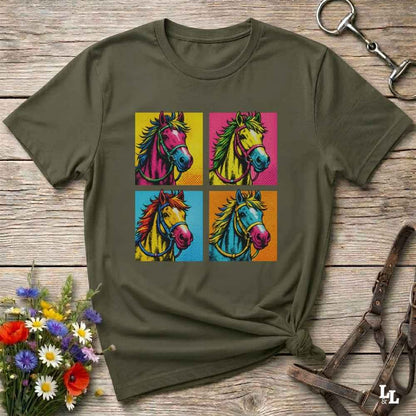 An olive green short-sleeve t-shirt with a colorful graphic print of four pop-art horse heads, styled on a rustic wooden background with equestrian accessories and flowers.