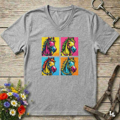 A heather gray V-neck t-shirt featuring a colorful pop-art graphic of four horse heads in a four-square grid. The shirt is displayed flat on a rustic wooden surface with equestrian tack and flowers as props.