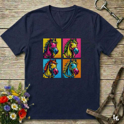 A navy blue V-neck t-shirt laid flat on a rustic wooden surface, featuring a colorful pop-art graphic of four horse heads in a grid. The shirt is styled with equestrian accessories, including a horse bit in the top right and a bridle strap in the bottom right, with a small bouquet of wildflowers in the bottom left.