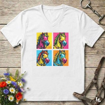 A white V-neck t-shirt laid flat on a rustic wooden surface, featuring a colorful graphic print of four stylized horse heads in a pop-art style. The t-shirt is styled with equestrian props, including a leather bridle and horse bits, and a small bouquet of wildflowers.