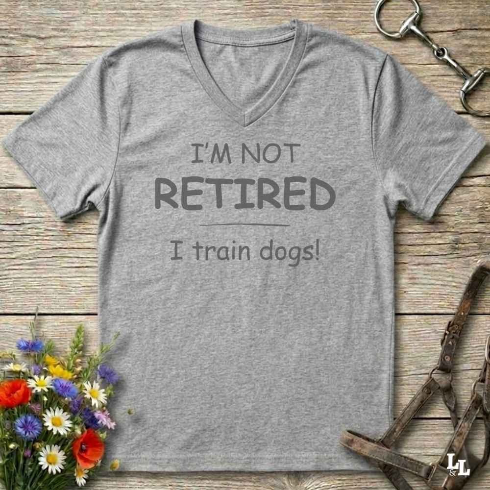 A heather gray V-neck t-shirt with the printed text 'I'M NOT RETIRED I train dogs!' in dark gray lettering, displayed on a rustic wooden background with decorative flowers, a horse bit, and a leather halter.