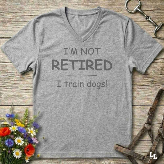 A heather gray V-neck t-shirt with the printed text 'I'M NOT RETIRED I train dogs!' in dark gray lettering, displayed on a rustic wooden background with decorative flowers, a horse bit, and a leather halter.