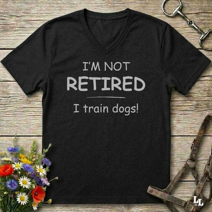 A black V-neck t-shirt with the text 'I'M NOT RETIRED I train dogs!' printed in white on the front, displayed on a rustic wooden background with dog harnesses, flowers, and a horse bit.