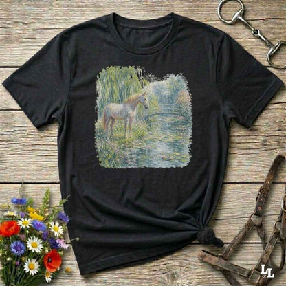 A black short-sleeve t-shirt with a square graphic print of a horse standing in a grassy field by a pond, laid flat on a rustic wooden surface with decorative flowers and horse tack around it.