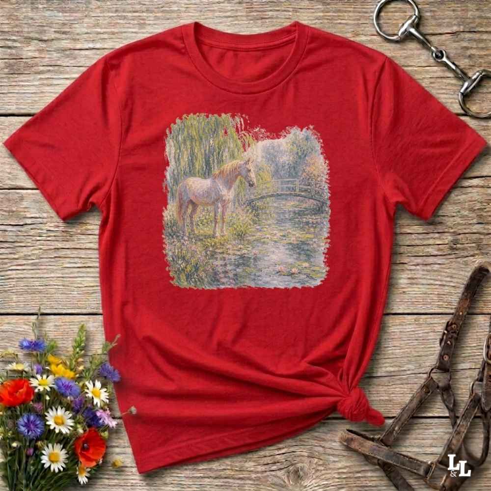 A red short-sleeve crewneck t-shirt with a square graphic print of a white horse standing in a grassy field by a pond. The shirt is styled in a flat-lay on a rustic wooden background with equestrian accessories and flowers.