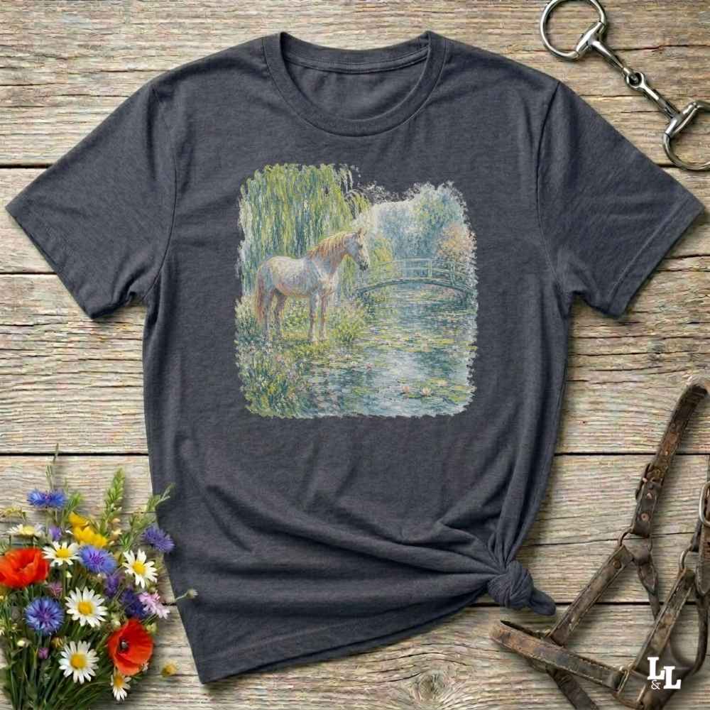 A dark charcoal gray t-shirt with a square graphic print of a white horse standing in a grassy pond, styled in a flat-lay on a rustic wooden background with flowers and horse tack.