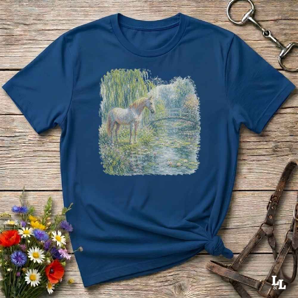 A navy blue short-sleeve t-shirt with a square graphic print of a white horse standing in a grassy field by a pond, styled on a rustic wooden background with equestrian accessories and flowers.