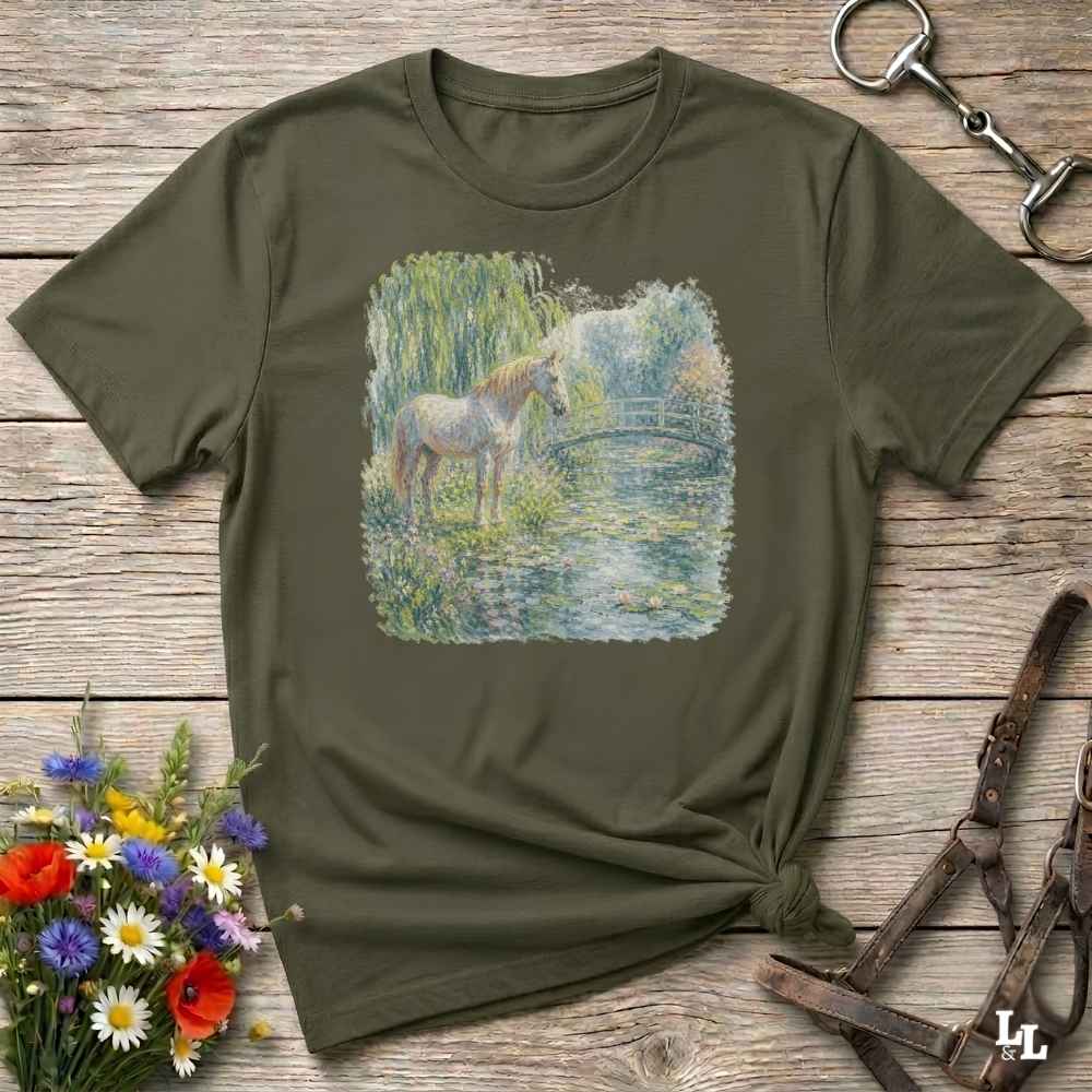 An olive green short-sleeve t-shirt with a square graphic print of a white horse standing in a pond, styled on a rustic wooden background with equestrian accessories and flowers.
