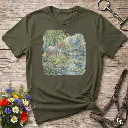 An olive green short-sleeve t-shirt with a square graphic print of a white horse standing in a pond, styled on a rustic wooden background with equestrian accessories and flowers.