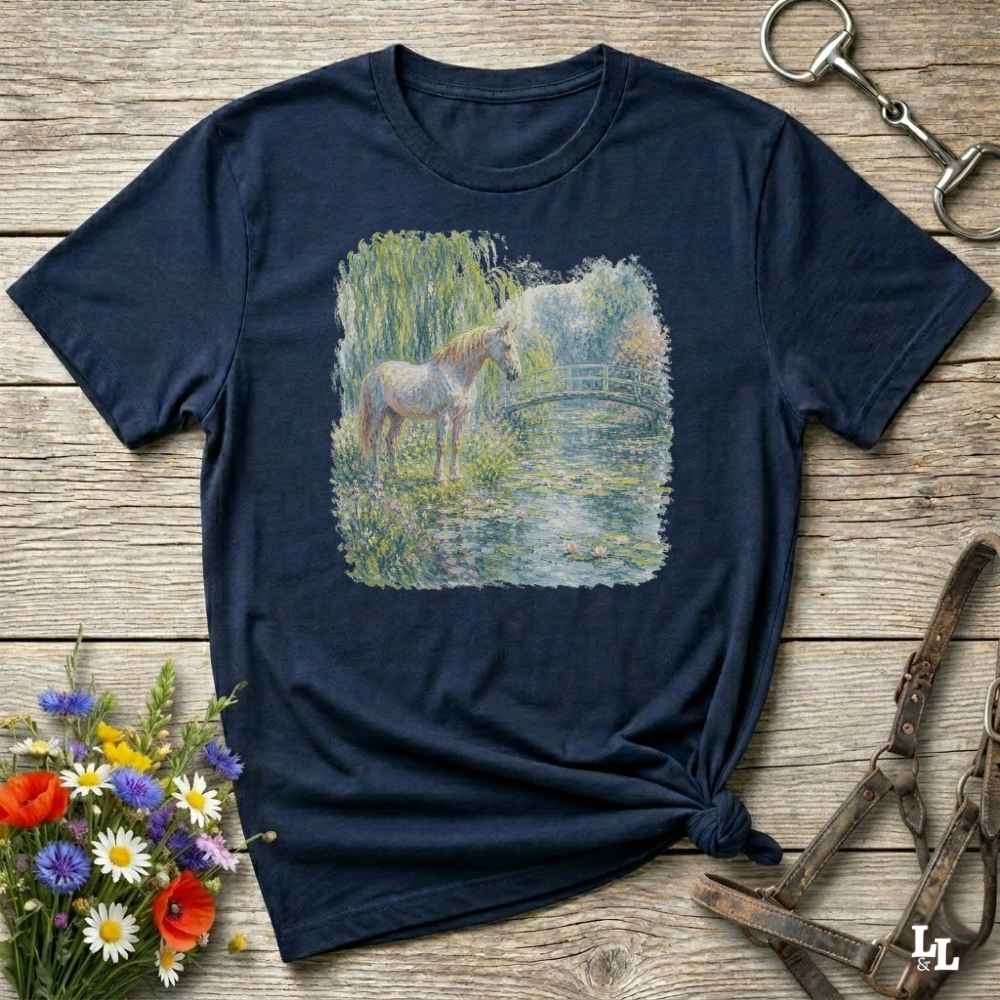 A navy blue short-sleeve t-shirt with a square graphic print of a horse standing in a grassy field by a pond, styled on a rustic wooden background with equestrian accessories and flowers.