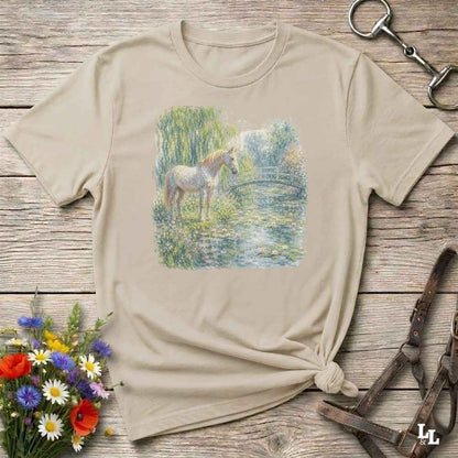 A beige short-sleeve t-shirt with a printed graphic of a white horse standing in a pond, styled on a rustic wooden background with equestrian accessories and a flower bouquet.