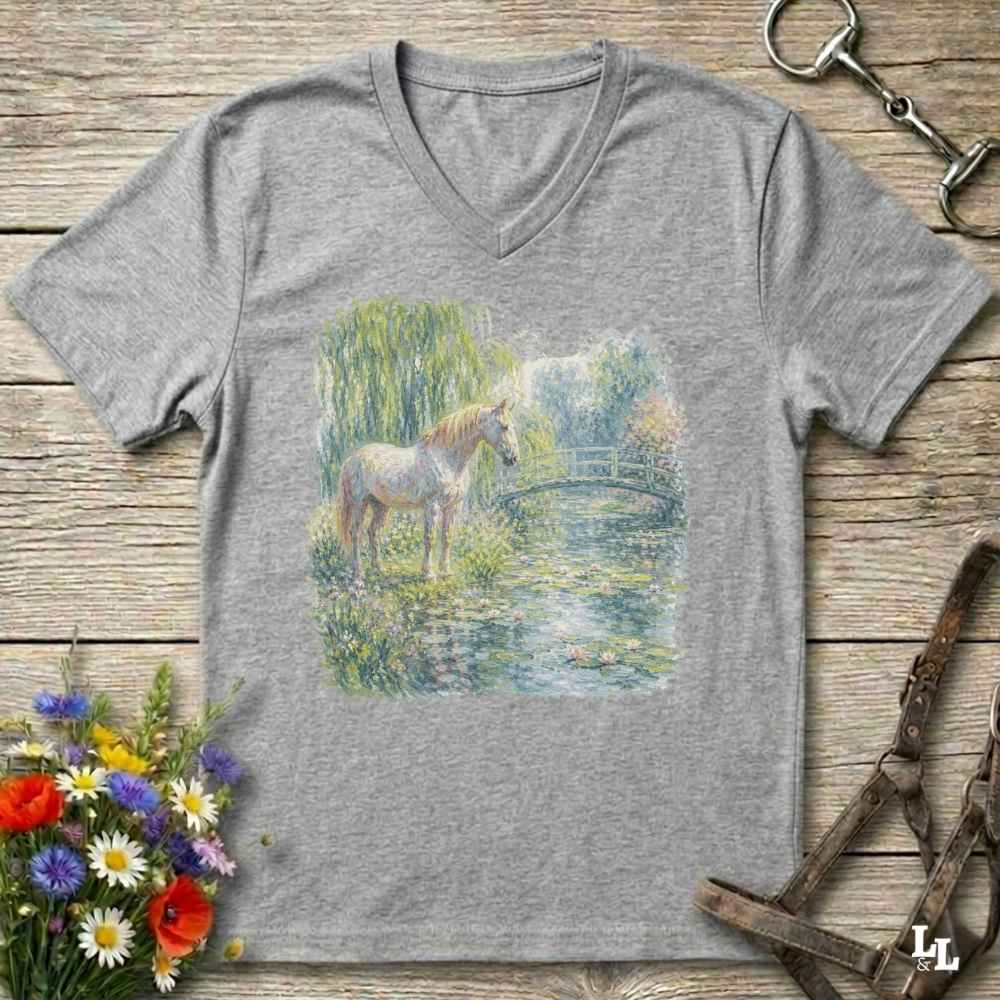 A grey V-neck t-shirt laid flat on a rustic wooden surface, featuring a colorful watercolor-style print of a horse standing by a pond. The shirt is styled with equestrian accessories (reins and bit) and a small bouquet of wildflowers.