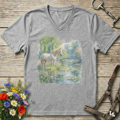A grey V-neck t-shirt laid flat on a rustic wooden surface, featuring a colorful watercolor-style print of a horse standing by a pond. The shirt is styled with equestrian accessories (reins and bit) and a small bouquet of wildflowers.