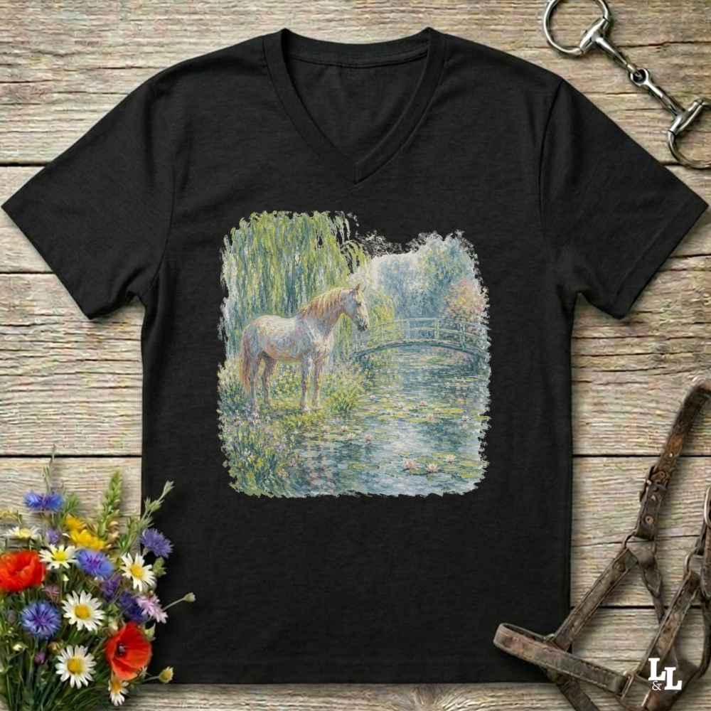 A black V-neck t-shirt laid flat on a rustic wooden surface, featuring a square graphic print of a white horse standing in a grassy pond. The shirt is styled with equestrian accessories, including a horse bit in the top right, a halter in the bottom right, and a bouquet of colorful wildflowers in the bottom left.