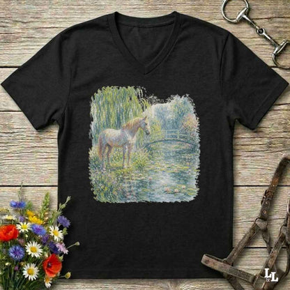 A black V-neck t-shirt laid flat on a rustic wooden surface, featuring a square graphic print of a white horse standing in a grassy pond. The shirt is styled with equestrian accessories, including a horse bit in the top right, a halter in the bottom right, and a bouquet of colorful wildflowers in the bottom left.