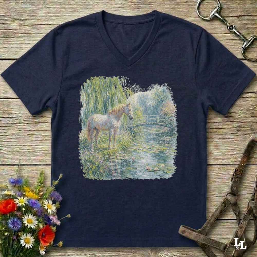 A navy blue V-neck t-shirt laid flat on a rustic wooden surface, featuring a central graphic print of a white horse standing in a grassy, water-filled landscape. The shirt is styled with equestrian accessories, including a halter, a horse bit, and a bouquet of colorful wildflowers.