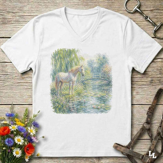 A white V-neck t-shirt laid flat on a rustic wooden surface, featuring a colorful watercolor-style print of a horse standing by a pond. The shirt is styled with equestrian accessories (reins and bit) and a small bouquet of wildflowers.