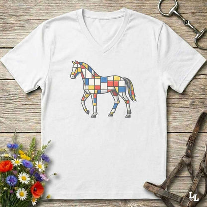 A white V-neck t-shirt laid flat on a rustic wooden surface, featuring a colorful geometric horse graphic on the chest. The shirt is styled with equestrian-themed props, including a horse bit in the top right, a halter in the bottom right, and a small bouquet of wildflowers in the bottom left.