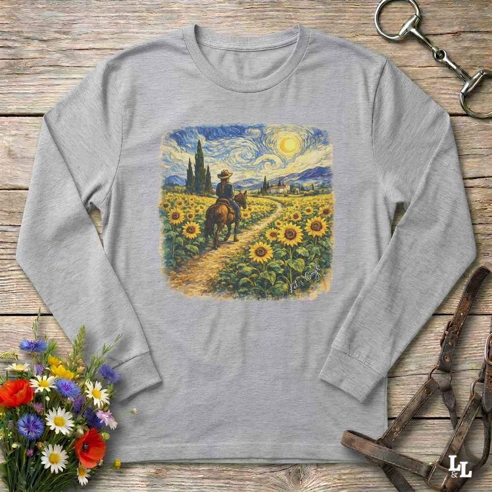 A light gray long-sleeve t-shirt with a square graphic print of a cowboy riding through a sunflower field in a Van Gogh-inspired style. The shirt is displayed flat on a rustic wooden background with decorative props including a bouquet of flowers and horse tack.