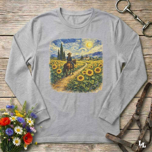 A light gray long-sleeve t-shirt with a square graphic print of a cowboy riding through a sunflower field in a Van Gogh-inspired style. The shirt is displayed flat on a rustic wooden background with decorative props including a bouquet of flowers and horse tack.