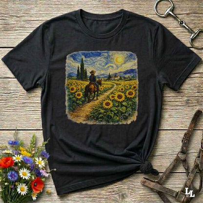 A black graphic t-shirt laid flat on a rustic wooden surface, styled with a bouquet of flowers, a horse bit, and a leather halter. The t-shirt features a square graphic print of a cowboy riding a horse through a sunflower field in a Van Gogh-inspired style.