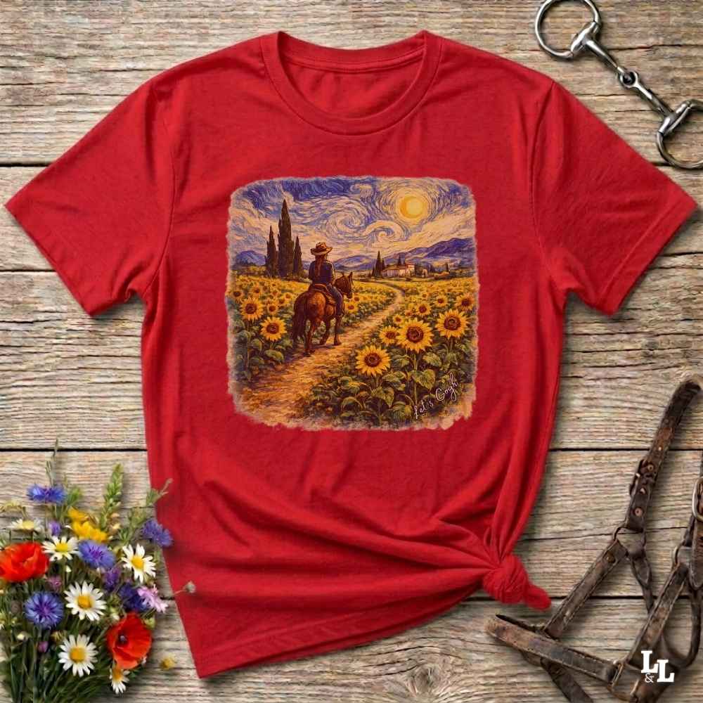 A red short-sleeve t-shirt with a square graphic print of a cowboy riding a horse through a sunflower field under a swirling starry sky. The shirt is styled flat on a rustic wooden background with decorative flowers and leather tack.
