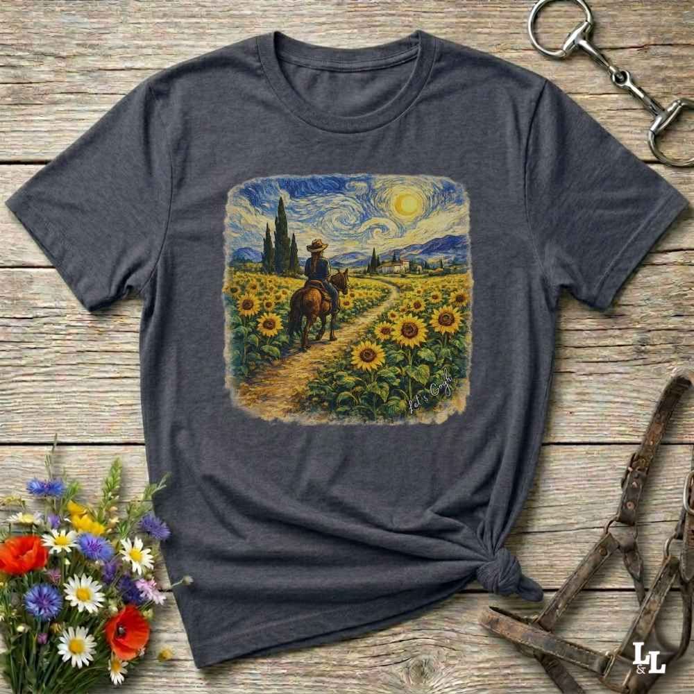 A dark gray heather t-shirt with a square graphic print of a cowboy riding through a sunflower field in a Van Gogh-inspired style. The shirt is displayed flat on a rustic wooden background with equestrian and floral props.